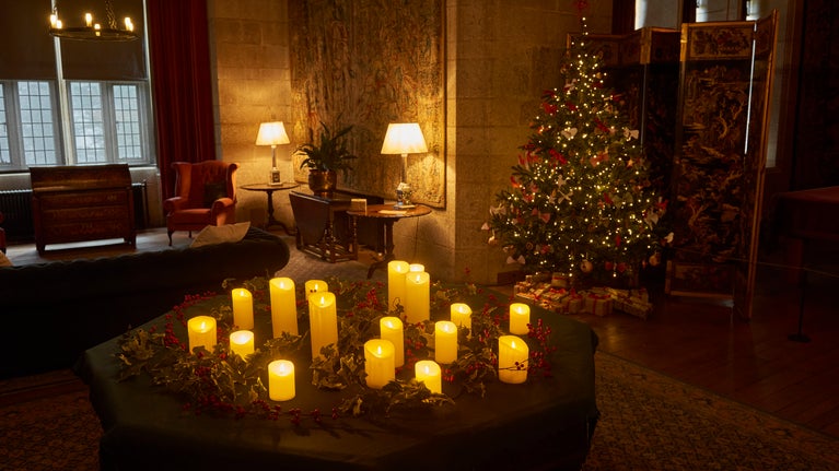 Father Christmas is sitting on a chair in a Library the room is dark with candles in the foreground and an old fashioned lamp in the background